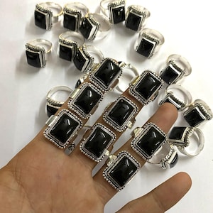 May include: Silver-toned rings with large, square, black gemstones. The rings have a detailed, beaded border around the stone. Some rings are on a hand, while others are scattered on a white surface. The rings are a bold, eye-catching accessory.