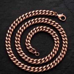 May include: A rose gold-toned curb chain necklace featuring interlocking flat, oval-shaped links. The necklace is arranged in a circular coil and includes a secure lobster claw clasp at one end and a small circular jump ring at the other end.