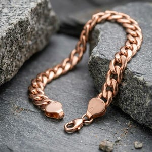 May include: A rose gold-toned curb chain bracelet featuring interlocking flat, oval-shaped links. The bracelet includes a secure lobster claw clasp mechanism at one end, with small, stamped hallmarks visible on the clasp components.