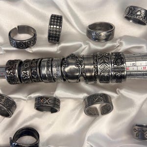 May include: A collection of silver rings with various designs, including floral patterns, geometric shapes, and a spoon-shaped ring. The rings are arranged on a white satin fabric background.