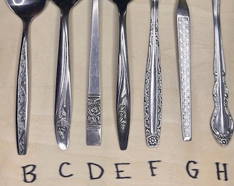 Op de afbeelding: Een verzameling van negen vintage zilveren lepels en serveerbestek, elk met unieke handvatontwerpen. De gebruiksvoorwerpen zijn gerangschikt op een lichtbruin houten oppervlak, met de letters A tot en met I.
