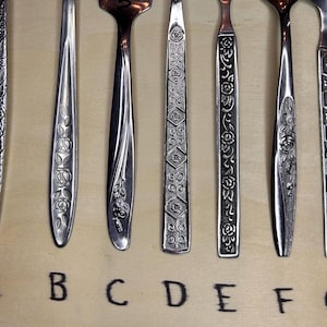 Pode incluir: Uma variedade de talheres vintage, colheres e garfos, com padrões florais e geométricos decorativos. As alças apresentam vários designs, incluindo rosas e gravuras detalhadas. As letras A a G estão escritas abaixo.