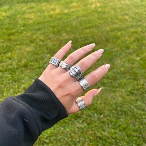 May include: A hand adorned with multiple silver rings featuring diverse designs, such as floral and geometric patterns. The rings are showcased against a background of green grass.