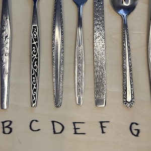 May include: A collection of nine vintage silver-toned flatware pieces, each with unique handle designs. The handles feature floral, scroll, and geometric patterns. Letters A through I are written below the flatware.