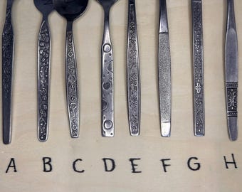 Op de afbeelding: Een assortiment vintage bestek, elk met unieke handvatontwerpen. De collectie omvat lepels, vorken en messen, met bloemen-, gestippelde en getextureerde patronen. Letters A tot en met H zijn onder het bestek gedrukt.