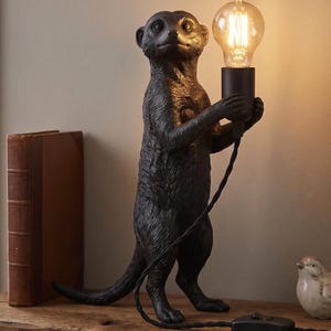 Könnte beinhalten: Eine schwarze Tischleuchte in Form eines stehenden Erdmännchens. Das Erdmännchen hält eine schwarze zylindrische Fassung mit einer leuchtenden, klaren Edison-Glühbirne. Ein schwarzes, gedrehtes Stoffkabel führt von der Fassung zu einem Schalter.