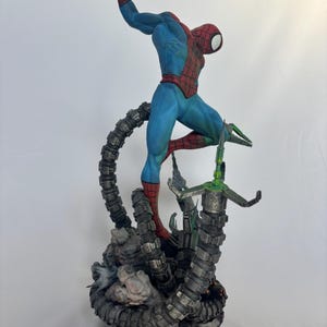 May include: A detailed figurine of Spider-Man in a dynamic pose, clad in a blue and red suit. The figure is atop a mechanical structure with metallic gray tentacles and green accents, set against a white backdrop. The statue is a collectible item.