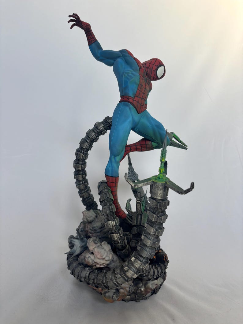 May include: A detailed figurine of Spider-Man in a dynamic pose, clad in a blue and red suit. The figure is atop a mechanical structure with metallic gray tentacles and green accents, set against a white backdrop. The statue is a collectible item.