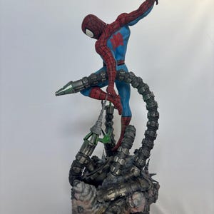 May include: A detailed figurine of Spider-Man in a dynamic pose, clad in a red and blue suit, with a web-like design on the mask. The figure is supported by mechanical tentacles, with a detailed base featuring a metallic and textured design.