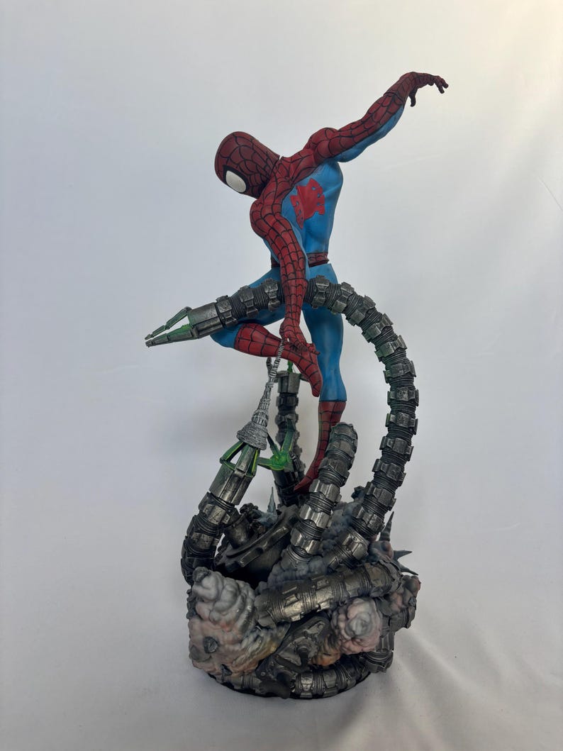 May include: A detailed figurine of Spider-Man in a dynamic pose, clad in a red and blue suit, with a web-like design on the mask. The figure is supported by mechanical tentacles, with a detailed base featuring a metallic and textured design.