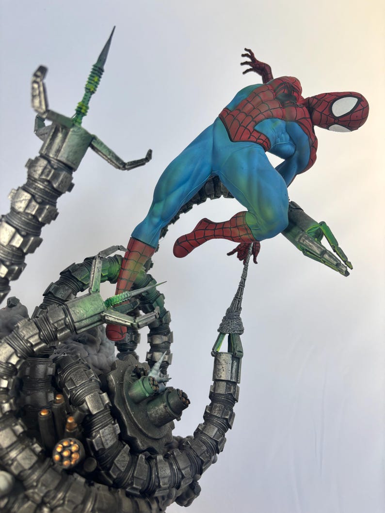 May include: A dynamic action figure of Spider-Man in a blue and red suit, posed mid-air, battling against Doctor Octopus's mechanical arms. The arms are metallic gray and green, with intricate details. The figure is set against a white background.