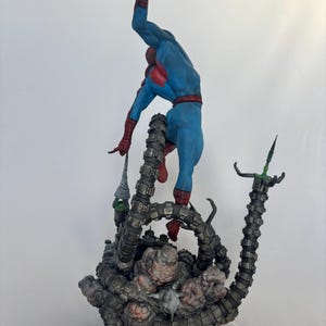 May include: A detailed figurine of Spider-Man in a dynamic pose, clad in a blue and red suit, seemingly battling against mechanical tentacles. The base features a detailed, textured design with a dark color palette.