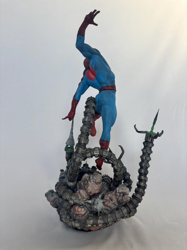 May include: A detailed figurine of Spider-Man in a dynamic pose, clad in a blue and red suit, seemingly battling against mechanical tentacles. The base features a detailed, textured design with a dark color palette.