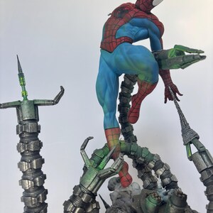 May include: A detailed figurine of Spider-Man in a dynamic pose, clad in his classic red and blue suit. He is perched atop a mechanical structure with multiple metallic green tentacles. The figure is set against a plain white background.