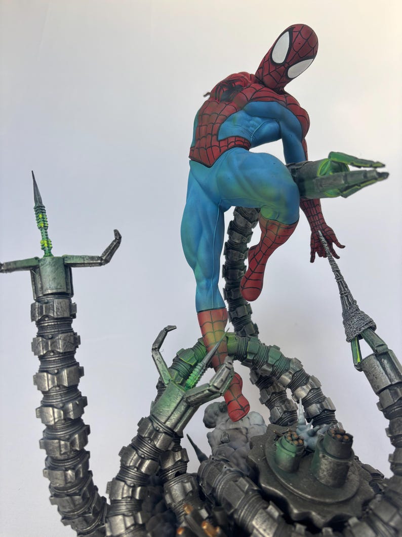 May include: A detailed figurine of Spider-Man in a dynamic pose, clad in his classic red and blue suit. He is perched atop a mechanical structure with multiple metallic green tentacles. The figure is set against a plain white background.
