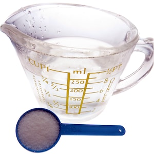 May include: A clear glass measuring jug with a handle, filled with water to the 100 ml mark. A small blue measuring spoon with white powder is next to the jug.