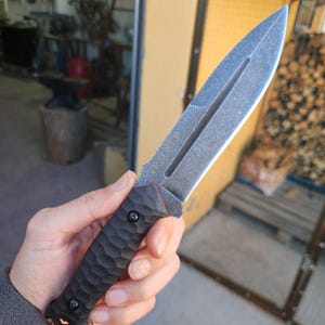May include: A fixed-blade knife with a textured, dark gray, hammered-finish handle secured by two black screws. The blade features a dark, weathered finish with a central fuller and the word TRACKER engraved near the hilt, ending in a sharp point.