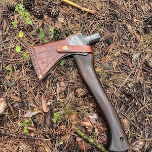 Puede incluir: Un hacha artesanal con mango de madera oscura y cabeza de metal, sobre un lecho de agujas y piñas de pino. El hacha tiene una funda de cuero marrón asegurada con un botón de latón. Herramienta para actividades al aire libre.