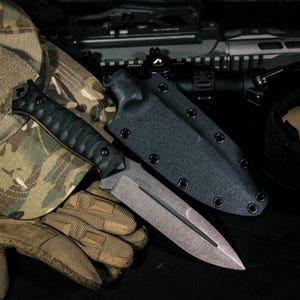 May include: A fixed-blade knife with a textured black handle and a double-edged steel blade featuring a central fuller. The knife is accompanied by a black molded sheath with metal eyelets, positioned alongside camouflage fabric and tan gloves.