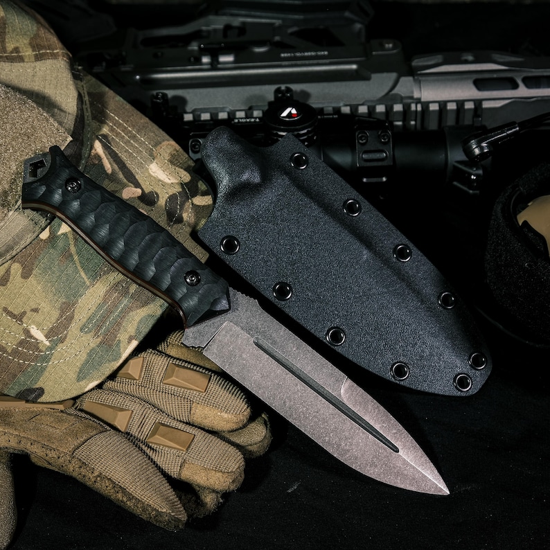 May include: A fixed-blade knife with a textured black handle and a double-edged steel blade featuring a central fuller. The knife is accompanied by a black molded sheath with metal eyelets, positioned alongside camouflage fabric and tan gloves.