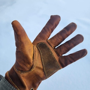 Könnte beinhalten: Ein brauner Lederhandschuh mit verstärkter Handfläche und Manschette. Der Handschuh hat sichtbare Nähte und eine kleine Metallöse in der Nähe des Handgelenks. Für Arbeiten oder Outdoor-Aktivitäten konzipiert.