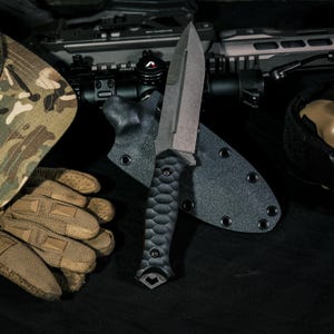 May include: A fixed-blade knife with a dark gray spear-point blade and a black textured handle featuring a scalloped grip pattern. The knife is positioned in front of a black molded sheath with metal eyelets, alongside tactical gear.