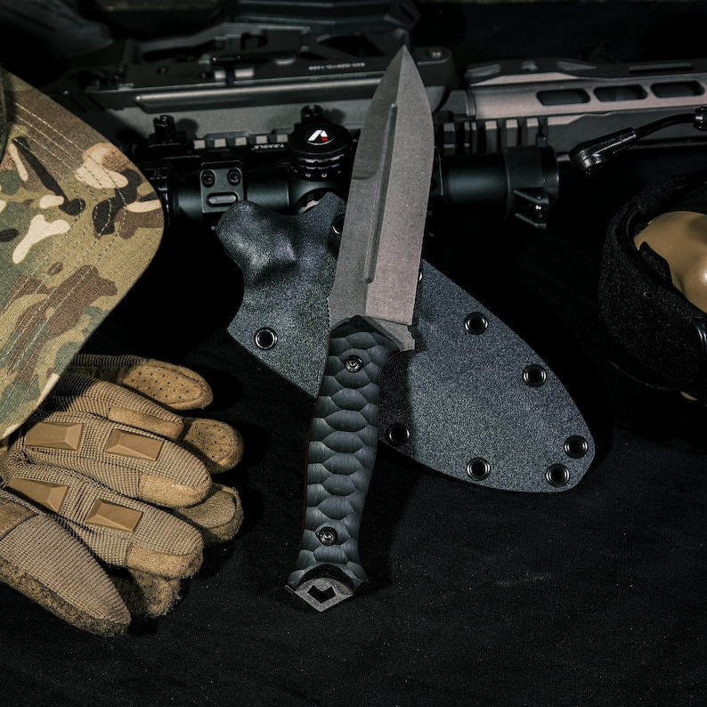 May include: A fixed-blade knife with a dark gray spear-point blade and a black textured handle featuring a scalloped grip pattern. The knife is positioned in front of a black molded sheath with metal eyelets, alongside tactical gear.