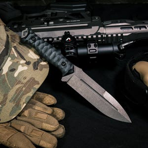 May include: A fixed-blade knife with a dark, textured handle and a matte gray steel blade featuring a central fuller. The knife is positioned alongside a camouflage patterned cap, tan tactical gloves, and a black rifle with an optical scope.