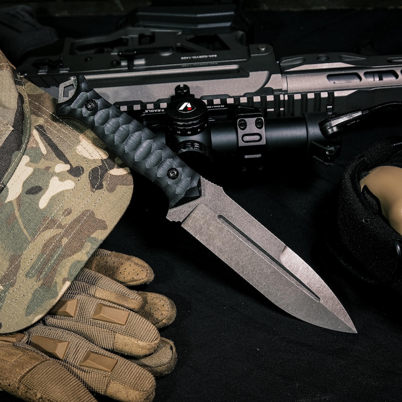 May include: A fixed-blade knife with a dark, textured handle and a matte gray steel blade featuring a central fuller. The knife is positioned alongside a camouflage patterned cap, tan tactical gloves, and a black rifle with an optical scope.