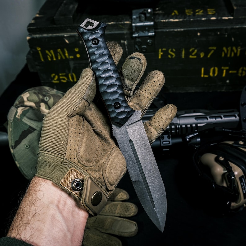 May include: A fixed-blade knife with a dark, textured, honeycomb-patterned handle and a matte gray steel blade featuring a central fuller groove. The handle is secured with two visible screws and has a distinct geometric pommel at the base.