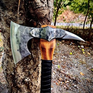 May include: A handcrafted axe with a polished steel blade and a wooden handle. The axe head has a curved blade and a pointed spike. The handle is wrapped in black leather for grip. The axe is leaning against a tree.