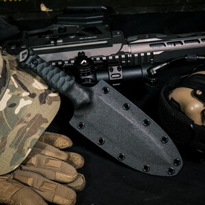 May include: A black knife with a textured handle and a matching black molded sheath featuring multiple metal eyelets along the edges. The sheath is designed for secure storage and protection of the blade, showcasing a functional and durable construction.