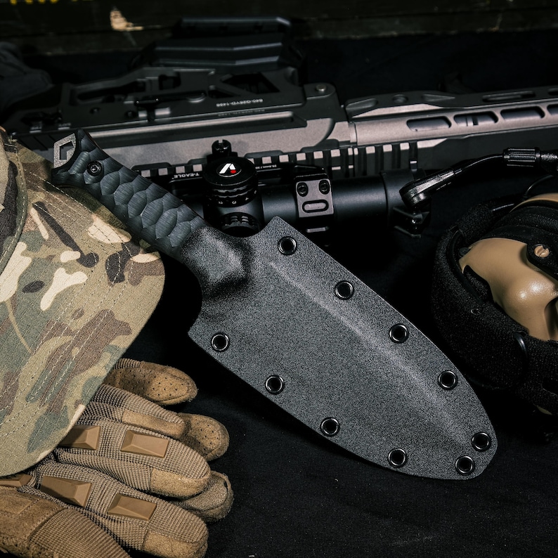 May include: A black knife with a textured handle and a matching black molded sheath featuring multiple metal eyelets along the edges. The sheath is designed for secure storage and protection of the blade, showcasing a functional and durable construction.