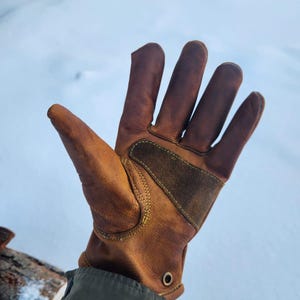 Può includere: Un guanto da lavoro in pelle marrone con una toppa in pelle scamosciata sul dorso della mano. Il guanto ha cuciture visibili e un occhiello metallico vicino al polso. Il guanto è usurato e mostra segni di utilizzo.