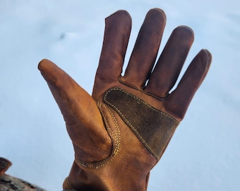 May include: A brown leather work glove with a suede patch on the back of the hand. The glove has visible stitching and a metal grommet near the wrist. The glove is worn and shows signs of use.