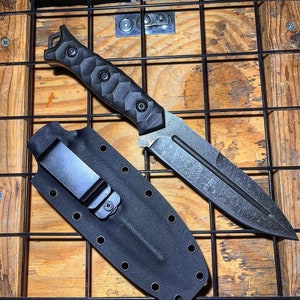 May include: A fixed-blade knife with a dark, textured finish and a black handle featuring a faceted, geometric grip pattern. The knife is accompanied by a black molded sheath with a metal belt clip and multiple eyelets along the edges.