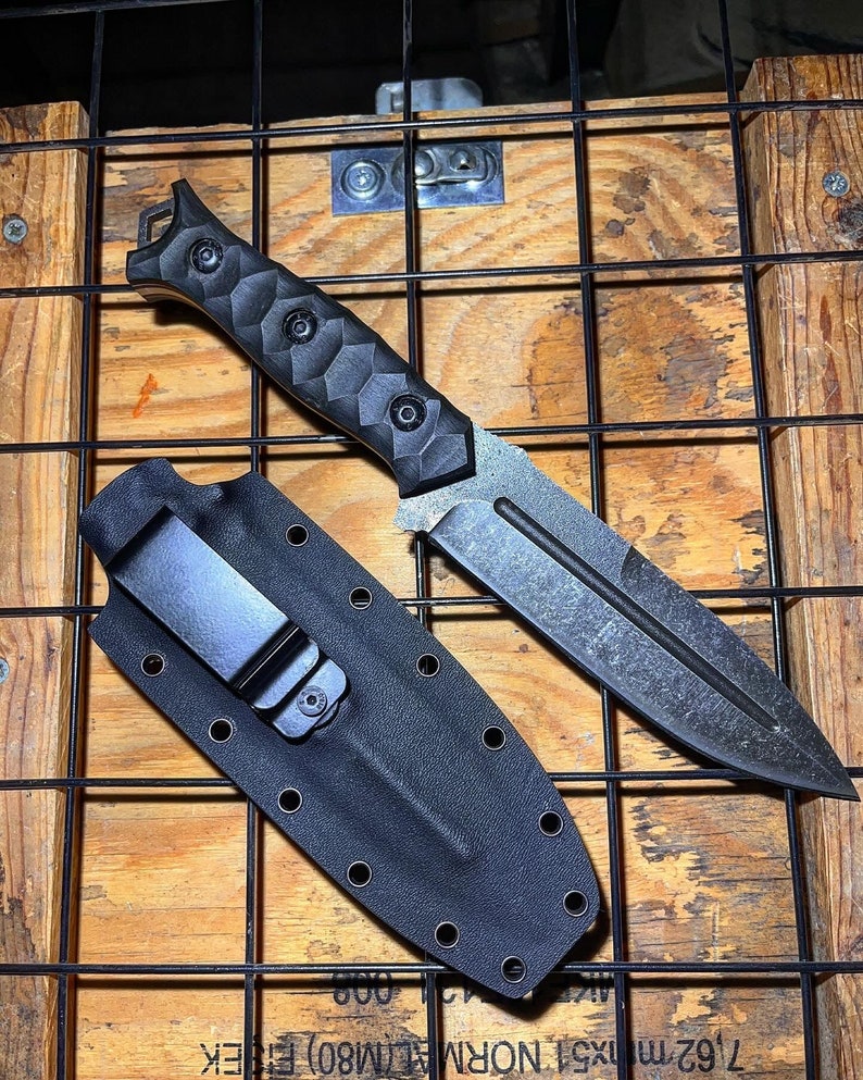 May include: A fixed-blade knife with a dark, textured finish and a black handle featuring a faceted, geometric grip pattern. The knife is accompanied by a black molded sheath with a metal belt clip and multiple eyelets along the edges.