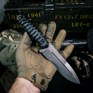May include: A fixed-blade knife with a dark, textured, ergonomic black handle and a matte grey steel blade featuring a central fuller groove. The knife is held in a tan tactical glove with perforated details and a metal snap button closure.