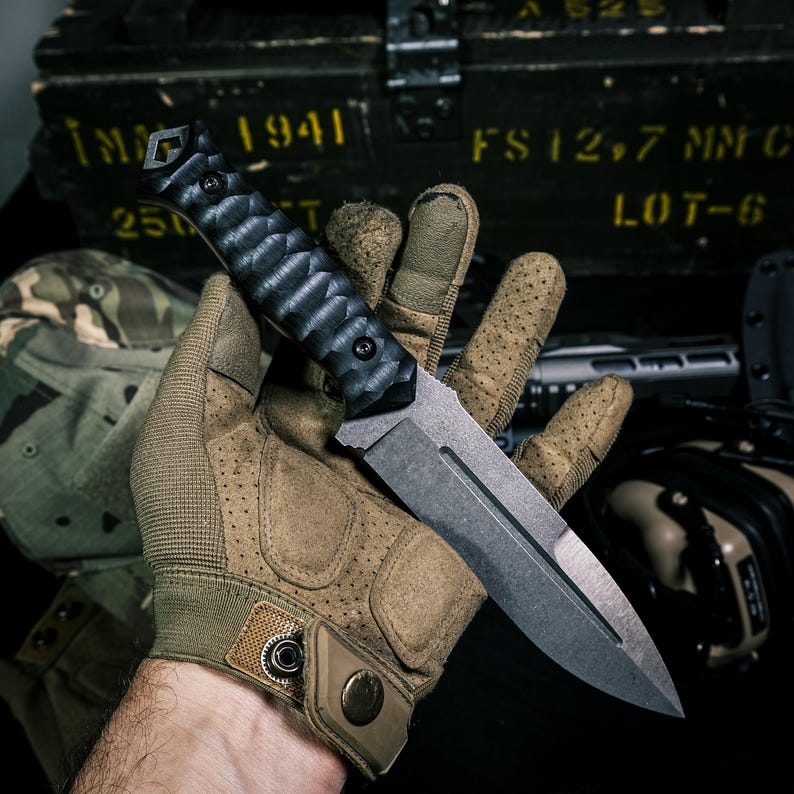 May include: A fixed-blade knife with a dark, textured, ergonomic black handle and a matte grey steel blade featuring a central fuller groove. The knife is held in a tan tactical glove with perforated details and a metal snap button closure.