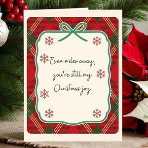 May include: Christmas card with a red and green plaid pattern border. The card features a cream-colored center with the text "Even miles away, you're still my Christmas joy" in black script, surrounded by snowflakes.