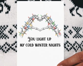 Könnte beinhalten: Eine weiße Grußkarte mit einem Herzmotiv aus Skeletthänden, verziert mit bunten Lichterketten. Auf der Karte steht in schwarzer Schrift "You light up my cold winter nights", ideal als Weihnachtsgeschenk.