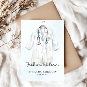 May include: A white coat ceremony announcement card featuring a watercolor illustration of a white lab coat on a hanger with a stethoscope draped over it. The text reads Joshua Wilson, WHITE COAT CEREMONY, and JUNE 23, 2027, in black font.