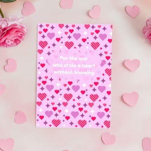 Op de afbeelding: Een roze wenskaart met een patroon van harten en sterren. De kaart heeft de tekst "For the one who stole a heart without knowing". Roze papieren harten en bloemen zijn rond de kaart verspreid.