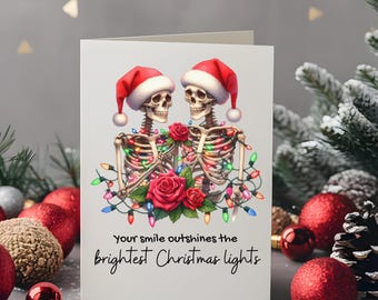 Könnte beinhalten: Weihnachtskarte mit zwei Skeletten mit Weihnachtsmützen, geschmückt mit Lichterketten und Rosen. Der Text auf der Karte lautet: "Your smile outshines the brightest Christmas lights."