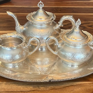 Könnte beinhalten: Ein silbernes Teeservice mit Teekanne, Milchkännchen, Zuckerdose und Tablett. Das Set hat ein verziertes Design mit Blumenmustern.