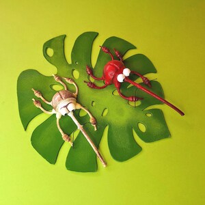 May include: Two whimsical, hand-painted, bug-shaped figurines with googly eyes. The red bug is on a green leaf-shaped cutout, while the beige bug is on the same cutout but with its long snout extending off the edge.