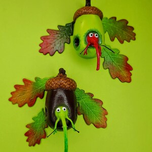 May include: Two acorn-shaped ornaments with whimsical faces and long noses. Each acorn is decorated with brown and green paint and has a pair of large, googly eyes. The acorns are attached to brown and green leaves.