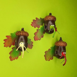 May include: Three acorn ornaments with painted faces and pipe cleaner noses. Each acorn is attached to a brown and green leaf.