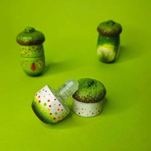 May include: Three acorn-shaped glass bottles with cork tops. The bottles are painted with green, brown, white, and yellow paint. One bottle is open and the glass dropper is visible.