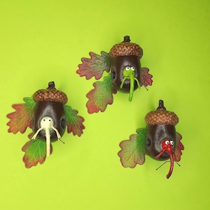 May include: Three acorn ornaments with painted faces and green, red, and white noses. Each acorn is attached to a brown leaf with red and green veins.